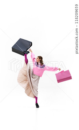Dressed in flowing pleated skirt and pink sweater, woman kicks high while holding shopping bags against white studio background. 118209659