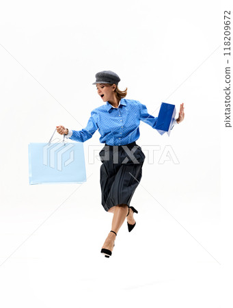 Leaping in joy with bags in hand, woman in blue polka-dot shirt and black skirt celebrates her shopping triumph against white studio background. 118209672