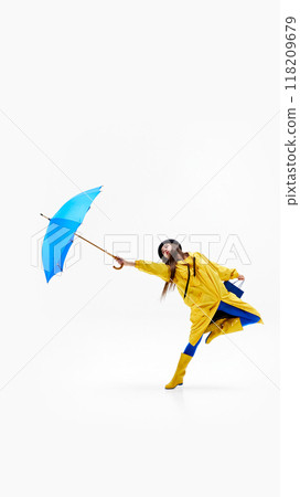 Cheerful woman in vibrant raincoat leaps with blue shopping bag and umbrella, embracing rainy weather against white background. 118209679