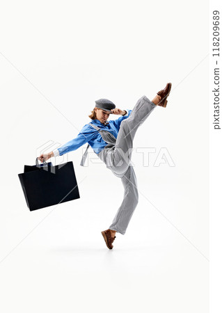 Young woman in grey overalls and blue shirt strikes playful pose, kicking one leg up and holding two shopping bags. 118209689
