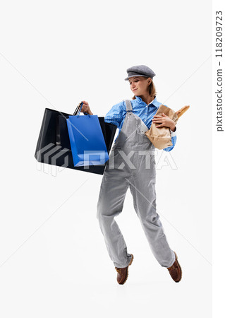 Dressed in casual overalls and cap, woman balances grocery and shopping bags in playful pose against white studio background. Dressed in casual overalls and cap, woman balances grocery and shopping bags in playful pose against white studio background. 118209723