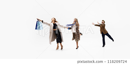 Women charge forward, shopping bags extended, while man trails behind in pursuit, against white studio background. 118209724