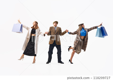 Trio dressed classical attire poses with shopping bags, holding hands in striking balance of style and energy against white studio background. Trio dressed classical attire poses with shopping bags, holding hands in striking balance of style and energy against white studio background. 118209727