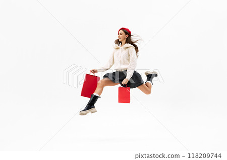 Stylish young woman, shopper leaping in motion, carrying red shopping bags against white studio background. Stylish young woman, shopper leaping in motion, carrying red shopping bags against white studio background. 118209744