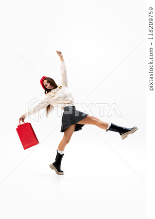 Jumping in joy, young fashionista, woman in red beret and cream sweater holds red shopping bag against white studio background. 118209759