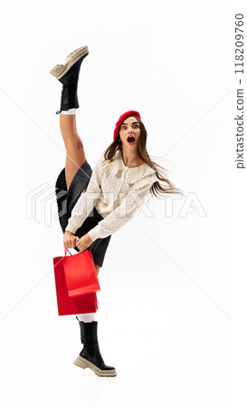 Young woman in sweater and black skirt holds red shopping bag while kicking her leg high, showcasing bold and playful style. 118209760