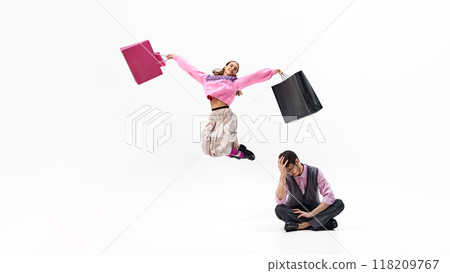 Woman leaps into air, filled with shopping joy, while her partner sits down in exhaustion against white studio background. 118209767