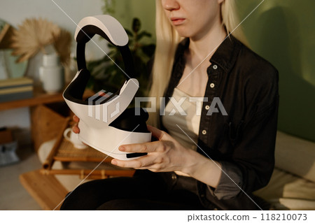 Medium section shot of young woman with long white hair holding modern VR headset in her hands while sitting on couch at home Medium section shot of young woman with long white hair holding modern VR headset in her hands while sitting on couch at home 118210073