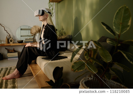 Side view wide shot of young girl with long white hair sitting on couch in wabi sabi living room watching movie via VR headset 118210085