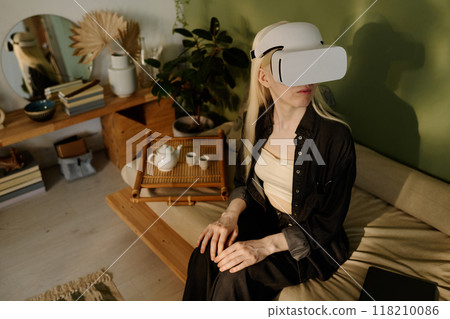 High angle view of young Caucasian woman with long white hair wearing VR headset sitting on couch in living room at home 118210086