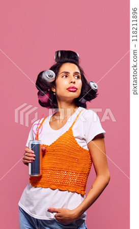 Woman looks away with playful expression, her hairstyle made of cans and drink in hand against pink background. Playful styling. Woman looks away with playful expression, her hairstyle made of cans and drink in hand against pink background. Playful styling. 118211896