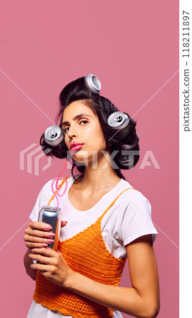 Young brunette woman with aluminum cans used as hair rollers holds drink with straw, posing against pink backdrop. Quirky fashion. Young brunette woman with aluminum cans used as hair rollers holds drink with straw, posing against pink backdrop. Quirky fashion. 118211897