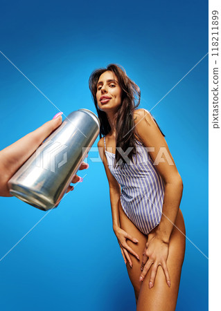 Young slim woman in swimsuit smiles as someone extends silver can toward her against bright blue backdrop. Summertime. Young slim woman in swimsuit smiles as someone extends silver can toward her against bright blue backdrop. Summertime. 118211899