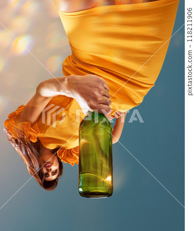 Young beautiful woman in sunny yellow dress holds large green bottle of sparkle wine posing against clear blue sky. Creative photo. 118211906