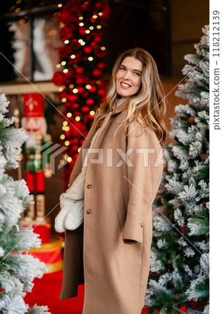 portrait of a happy woman in a coat and white mittens posing against the backdrop of Christmas decorations, the mood of the approaching holiday 118212139
