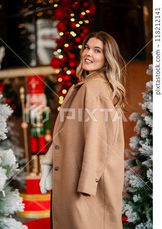 portrait of a happy woman in a coat and white mittens posing against the backdrop of Christmas decorations, the mood of the approaching holiday portrait of a happy woman in a coat and white mittens posing against the backdrop of Christmas decorations, the mood of the approaching holiday 118212141