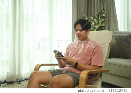 Smiling young asian man using mobile phone on armchair at home Smiling young asian man using mobile phone on armchair at home 118212681