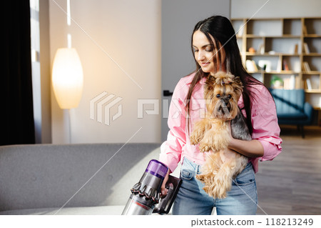 Woman with vacuum cleaner and dog in her hand vacuuming in the living room. Woman with vacuum cleaner and dog in her hand vacuuming in the living room. 118213249