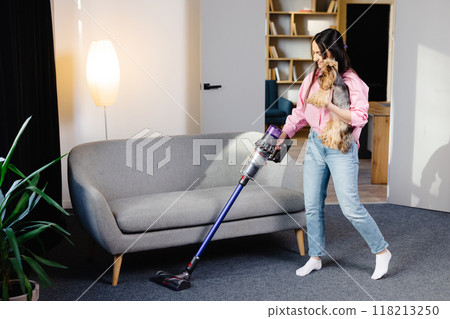 Woman with vacuum cleaner and dog in her hand vacuuming in the living room. Woman with vacuum cleaner and dog in her hand vacuuming in the living room. 118213250