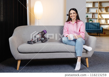 Young woman sitting on sofa next to cordless vacuum cleaner at home and holding cup in hands. Housework and chores concept. Cleaning appliances Young woman sitting on sofa next to cordless vacuum cleaner at home and holding cup in hands. Housework and chores concept. Cleaning appliances 118213259
