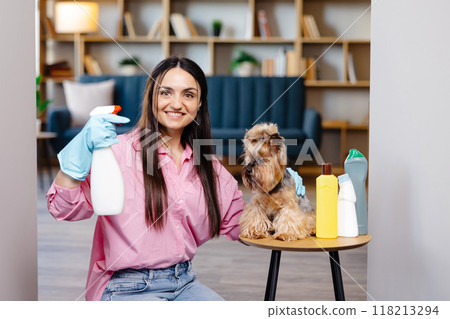 Young woman sitting near the table with cleaning products at home and her little dog is sitting on the table too 118213294