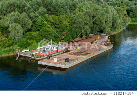 Docked barge and small boat by a green forested riverside. 118214194