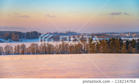 A serene winter landscape unfolds at sunset, showcasing expansive snow-covered fields under a pastel sky. The distant trees stand bare against the fading light, creating a tranquil atmosphere. 118214768