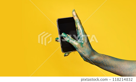 Hand covered in green slime holding black smartphone with blank screen against yellow background. Halloween-themed image for social media and marketing materials. 118214832