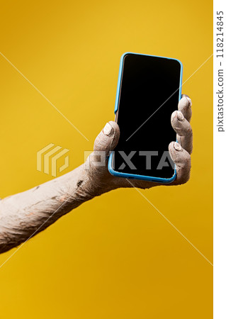 Zombie-like hand with long, dirty fingernails holding mobile phone with blank screen against yellow background. News, sales, announcement 118214845