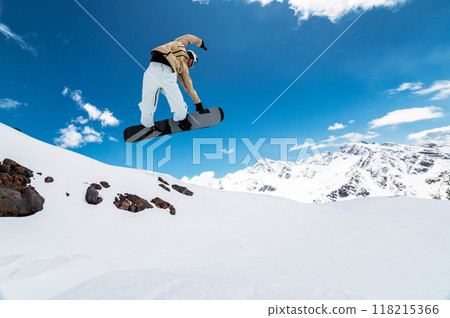 snowboarder jumps from a big air with a board grab, a nose or tail trick in rotation, against the backdrop of snow-capped mountains and the bright sun 118215366