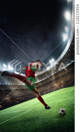 Under floodlights on stadium, soccer player in red and green kits unleashes powerful kick toward the goal, poised to make an unforgettable strike in final game. Under floodlights on stadium, soccer player in red and green kits unleashes powerful kick toward the goal, poised to make an unforgettable strike in final game. 118215954