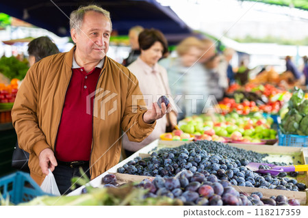 Positive mature male holding plum while choosing fruits and vegetables at market Positive mature male holding plum while choosing fruits and vegetables at market 118217359