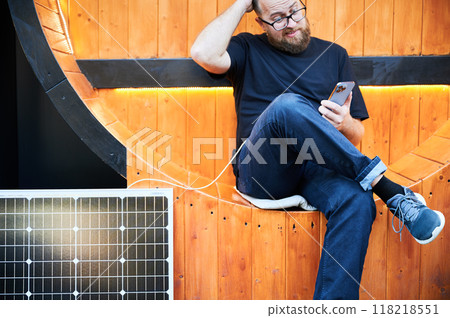Bearded man using smartphone connected to photovoltaic solar panel. Integration of sustainable renewable energy into everyday life, showing practical use of solar power. Cropped view. Focus on phone. 118218551