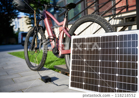 Pink electric mountain bike parked beside staircase, connected to small solar panel for charging. Bike rugged tires, mounted helmet on handlebar, situated on paved pathway with grassy lawn. Pink electric mountain bike parked beside staircase, connected to small solar panel for charging. Bike rugged tires, mounted helmet on handlebar, situated on paved pathway with grassy lawn. 118218552