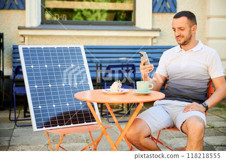 Young man charging smartphone from solar panel while sitting in cafe. Handsome male drinking coffee and checking his phone next to solar battery. Conscious male choosing sustainable lifestyle. 118218555