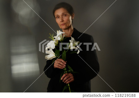 Waist up portrait of senior woman in black elegant suit holding white lilies grieving loss, while looking seriously at camera with stare expressing sadness on gray background in studio, copy space 118219188