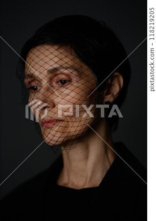 Vertical portrait with deep shadows of pensive senior woman wearing headband with mesh mourning veil, looking down in silent grief on black background in dim studio 118219205