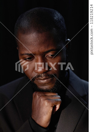 Vertical portrait of pensive Black man in formal suit looking down posing with hands near face in low light on dark background in studio Vertical portrait of pensive Black man in formal suit looking down posing with hands near face in low light on dark background in studio 118219214