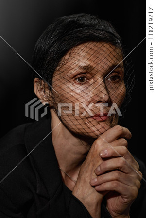 Portrait of pensive senior woman wearing headband with mesh mourning veil posing with hands near face expressing silent grief on black background in studio Portrait of pensive senior woman wearing headband with mesh mourning veil posing with hands near face expressing silent grief on black background in studio 118219217