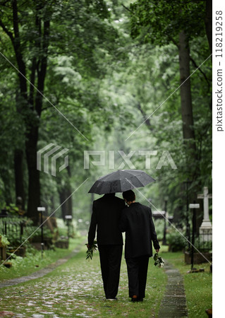 Vertical shot of grieving couple in black suits walking under umbrella along paved alley at cemetery featuring tall old trees with lively greenery, copy space Vertical shot of grieving couple in black suits walking under umbrella along paved alley at cemetery featuring tall old trees with lively greenery, copy space 118219258