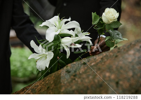 Close up on rain wet white lilies and rose in hands of two unrecognizable people placing flowers on stone memorial monument to commemorate deceased loved one at cemetery, copy space 118219288