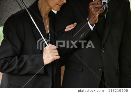 Cropped shot of senior woman in black suit holding husband by arm while hiding from rain under umbrella, copy space Cropped shot of senior woman in black suit holding husband by arm while hiding from rain under umbrella, copy space 118219306