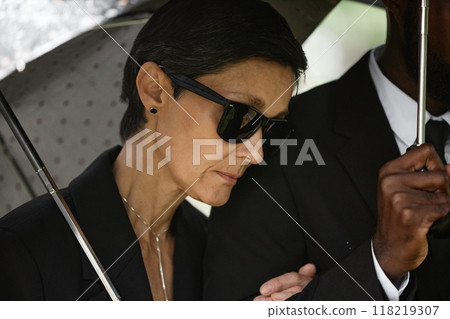 Close up of grieving senior woman in black sunglasses standing by husband holding supportive man by arm, while hiding from rain under umbrella at memorial service at cemetery 118219307