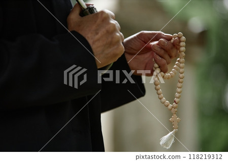 Close up on hands of unrecognizable senior person holding wooden rosary beads praying for soul of deceased at gravesite while hiding from rain under umbrella, copy space 118219312