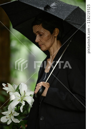 Vertical shot of senior woman in black suit and mourning veil under umbrella grieving over death of loved one at cemetery, holding white lilies wet from rain before placing flowers at grave 118219325