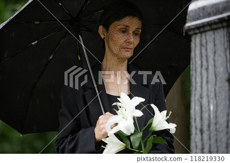 Medium shot of senior woman in mourning veil holding white lilies visiting grave of passed away loved one, while hiding from rain under umbrella at cemetery Medium shot of senior woman in mourning veil holding white lilies visiting grave of passed away loved one, while hiding from rain under umbrella at cemetery 118219330