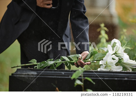 Close up on hand of unrecognizable senior woman placing white lilies on tombstone in memory of departed loved one while raindrops falling on grave at cemetery, copy space Close up on hand of unrecognizable senior woman placing white lilies on tombstone in memory of departed loved one while raindrops falling on grave at cemetery, copy space 118219331