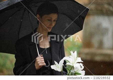 Medium shot of sorrowful senior woman in black funeral suit and veil mourning loss of loved one hiding from rain under umbrella before placing white lilies on grave at cemetery, copy space Medium shot of sorrowful senior woman in black funeral suit and veil mourning loss of loved one hiding from rain under umbrella before placing white lilies on grave at cemetery, copy space 118219332