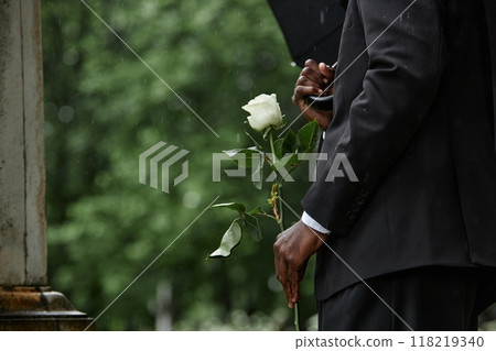 Cropped shot of Black man in funeral black suit holding white rose in memory of deceased loved one before placing flower on grave in rainy cemetery, copy space Cropped shot of Black man in funeral black suit holding white rose in memory of deceased loved one before placing flower on grave in rainy cemetery, copy space 118219340