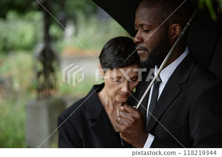 Side view of sorrowful Black man comforting grieving senior woman leaning on his shoulder at memorial service. Mourning couple standing under umbrella at cemetery, copy space 118219344